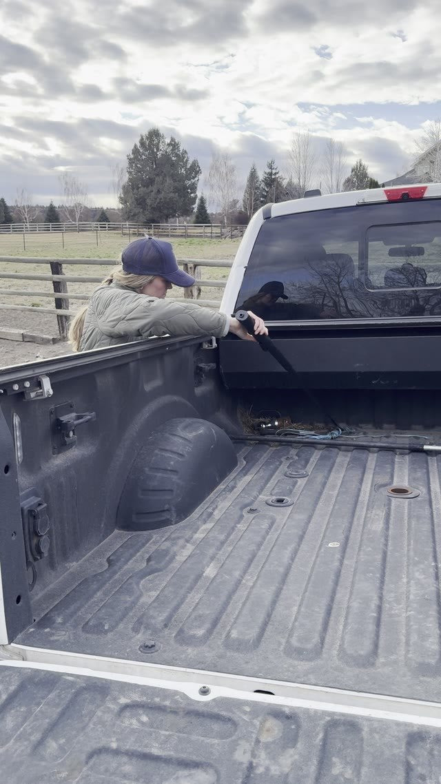 A person in a cap leans over the side of a pickup truck bed and reaches in with the extended Lazy Cowboy hook.